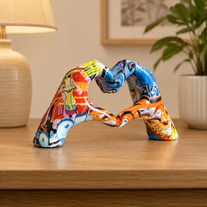 Può includere: Una scultura decorativa di due mani che formano un cuore, ricoperta da un vivace motivo in stile graffiti multicolore con sfumature di blu, arancione, rosso e giallo, caratterizzata da forme e linee astratte su tutta la superficie.