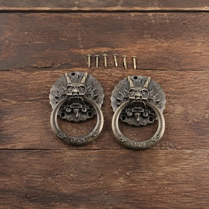 May include: Two antique bronze door knockers with a dragon design, each with a circular handle. The knockers are mounted on a wooden surface, with several screws above them. The knockers have a vintage, ornate appearance.
