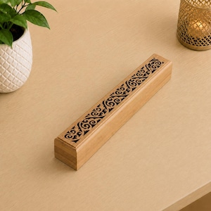 Puede incluir: Una caja rectangular de madera para incienso con un acabado marrón claro. La tapa superior presenta un intrincado patrón de volutas cortado con láser que revela un interior oscuro. La caja tiene una forma larga y delgada con bordes lisos.