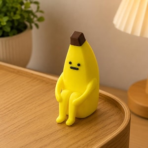 Op de afbeelding: Een geel, banaanvormig figuurtje met een bruine bovenkant, voorzien van een eenvoudig gezicht met zwarte stippen en een streepje. Het figuurtje zit in een zittende houding met de armen om de knieën geslagen en heeft een glad, compact ontwerp.