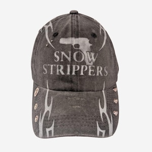 Könnte beinhalten: Eine anthrazitfarbene Baseballkappe im Used-Look mit einer weißen Grafik einer Pistolensilhouette über dem Schriftzug SNOW STRIPPERS. Die Kappe ist mit weißen Tribal-Flammenmustern auf der Vorderseite und an den Seiten sowie silbernen Nieten versehen.