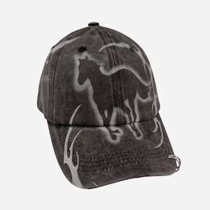 Puede incluir: Una gorra de béisbol de color gris carbón lavado con una silueta de caballo aerografiada en gris claro en los paneles frontales y patrones de llamas en la visera. Un pequeño anillo de metal plateado está sujeto al lado derecho de la visera.
