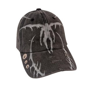 Può includere: Cappellino da baseball nero con un aspetto invecchiato, caratterizzato da una grafica grigio chiaro di una creatura alata. Il cappellino ha occhielli metallici e rivetti decorativi. Un accessorio alla moda.