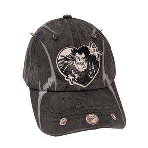Könnte beinhalten: Eine verwaschene, anthrazitfarbene Baseballkappe mit silbernen Metallspitzen und Ösen. Auf der Vorderseite befindet sich ein herzförmiger Aufnäher mit einer Schwarz-Weiß-Illustration einer Figur. Die Kappe hat eine abgenutzte, Vintage-Ästhetik.