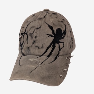 May include: A distressed grey baseball cap featuring a black airbrushed spider graphic on the front panel. The cap is adorned with silver-toned metal spikes along the side and brim, along with small metal studs and decorative stitching details.