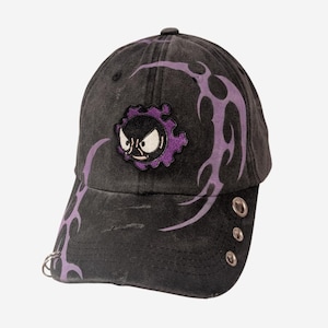 Può includere: Un cappellino da baseball nero dall'aspetto vissuto, caratterizzato da un ricamo viola che raffigura un personaggio stilizzato con un'espressione arrabbiata. Il cappellino è decorato con motivi a fiamma tribali viola e tre occhielli metallici argentati.