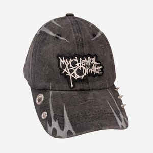 Puede incluir: Una gorra de béisbol gris carbón con efecto desgastado que presenta un parche blanco con el logotipo de My Chemical Romance en la parte frontal. Incluye ojales metálicos plateados, pinchos plateados en la visera y un estampado de llamas.