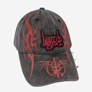 Può includere: Un cappellino da baseball grigio antracite con grafiche a fiamme tribali rosse sui lati e sulla visiera. La parte anteriore presenta una toppa ricamata con il logo rosso Lamb of God, con una grafica circolare rossa sottostante. Borchie in metallo argentato sporgono dal lato.