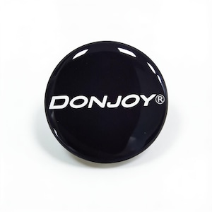 May include: A circular, glossy black button or emblem featuring the white text DONJOY followed by a registered trademark symbol. The surface is smooth and reflective, with a clean, rounded edge and a bold, high-contrast design.