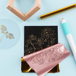 May include: A black card with a gold floral foil design, a sheet of rose gold foil, a gold foil roll, a white and teal craft pen, a clear circular disc with a gold dragonfly print, and a wooden frame piece on a light blue surface.