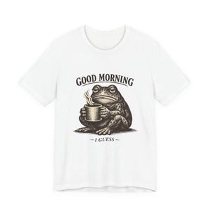 May include: White t-shirt featuring a grumpy frog holding a coffee cup. The frog illustration is in a vintage style with the words "GOOD MORNING - I GUESS -" above and below the frog. The shirt is a casual wear item.