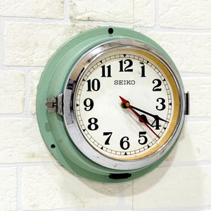 Puede incluir: Un reloj de pared redondo Seiko con carcasa de metal verde pálido y bisel de cromo pulido. La esfera es blanca con números arábigos negros, manecillas de hora y minutos negras y un segundero rojo fino. El nombre de la marca Seiko está impreso.