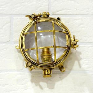 Op de afbeelding: Een ronde, messingkleurige nautische wandlamp met een heldere, met raster bedekte glazen lens. De armatuur heeft een lamphouder en is gemonteerd op een witte bakstenen muur. De lamp heeft een vintage, industriële uitstraling.