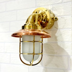 May include: A nautical-style wall sconce featuring a polished brass mounting base and curved arm. The fixture includes a wide copper shade and a cylindrical frosted glass diffuser protected by a brass wire cage, creating a robust industrial look.
