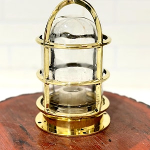 May include: A polished brass nautical bulkhead light fixture featuring a cylindrical clear glass dome protected by a metal cage frame with a top handle and a circular base with pre-drilled mounting holes and a central white ceramic socket.