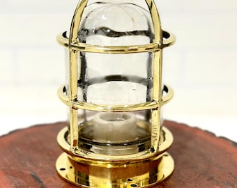 May include: A polished brass nautical bulkhead light fixture featuring a cylindrical clear glass dome protected by a metal cage frame with a top handle and a circular base with pre-drilled mounting holes and a central white ceramic socket.