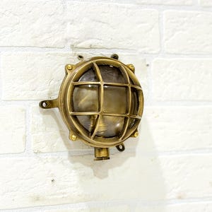 May include: A round nautical-style bulkhead wall light fixture made of polished brass. The circular frame features a protective metal cage over a clear glass lens, secured by small screws around the perimeter, with a small cylindrical base attachment.