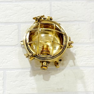 Puede incluir: Aplique de pared redondo de estilo náutico en color latón. La lámpara tiene un diseño en forma de jaula con una carcasa de vidrio y un portalámparas. El aplique está montado en una pared de ladrillo blanco.