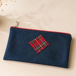 May include: A rectangular dark blue denim zipper pouch featuring a red, green, and white plaid fabric patch stitched in a diamond orientation in the center. The pouch has a bright red zipper tape and a silver metal zipper pull.
