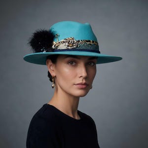Könnte beinhalten: Ein leuchtend türkisfarbener Fedora-Hut mit breiter Krempe und einem dekorativen Band. Das Band besteht aus einem gemusterten Stoffstreifen, einer dunkelblauen Kordel, einem schwarzen Feder-Pompon und einem kleinen goldfarbenen Flamingo-Anhänger.