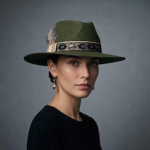 Könnte beinhalten: Ein olivgrüner Fedora-Hut mit breiter Krempe und strukturierter Krone. Der Hut ist mit einem beigen Band verziert, das mit einer schwarzen floralen Spitzenborte überlagert ist, sowie einem kleinen Büschel grauer und brauner Federn.