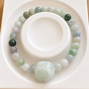 May include: A beaded bracelet featuring smooth, round stones in varying shades of translucent green, white, and soft lavender. A larger, carved stone bead with a floral-like pattern serves as the central focal point of the circular jewelry piece.