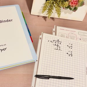 May include: A light blue binder labeled Science Binder Property of Harper with colorful index tabs, a sheet of graph paper with handwritten long division math problems, a black pen, and a printed flowchart page on a light wooden surface.