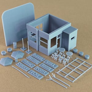 May include: A light gray 3D printed miniature building kit featuring a small structure, tables, chairs, umbrellas, and rectangular signs with the text Döner, Curry, Ecke, Imbiss, Bistro, Grill, Kiosk, and Point, all rendered in a matte finish.