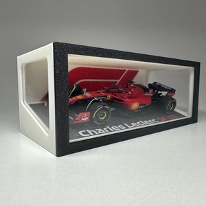 Può includere: Un modello di auto da corsa di Formula 1 rosso e nero all'interno di una vetrina. L'auto ha il numero 16 e i loghi degli sponsor. La vetrina ha una cornice nera e il testo "Charles Leclerc SF-75".