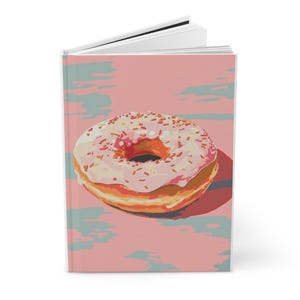 May include: A spiral-bound notebook featuring a colorful illustration of a glazed donut with sprinkles. The donut is set against a pink background with blue and gray brushstrokes. The notebook's cover is a vibrant and eye-catching design.