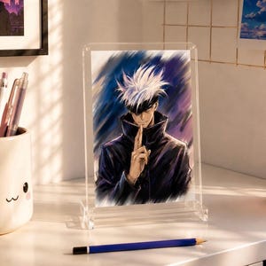 May include: A clear acrylic frame holding a vertical art print of an anime character with spiky white hair and a black blindfold. The character wears a dark high-collared jacket and holds one finger to their lips in a gesture of silence.