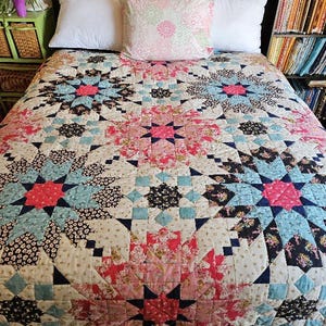 Op de afbeelding: Een sprei met een herhalend sterpatroon in tinten roze, lichtblauw, crème en zwart. Het ontwerp bestaat uit complexe geometrische patchworkblokken met bloemmotieven en kleinschalige prints, wat zorgt voor een symmetrische indeling.
