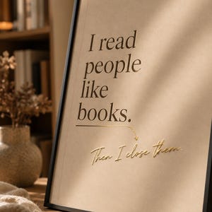 Könnte beinhalten: Ein gerahmter Kunstdruck mit hellbraunem Hintergrund und schwarzem Text, der lautet: I read people like books. Darunter zeigt ein goldener Pfeil auf den kursiven goldenen Text: Then I close them. Der Rahmen ist dünn, schwarz und schräg aufgestellt.