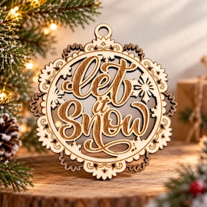 Puede incluir: Un adorno circular de madera con un diseño cortado a láser que incluye el texto "let it Snow" en el centro. El adorno presenta un borde festoneado intrincado con patrones de copos de nieve y un pequeño orificio en la parte superior.