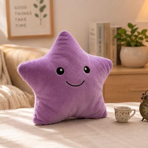 May include: A plush, lavender star-shaped pillow with a smiling face and black eyes. The pillow is on a bed with a small teacup and a vintage alarm clock. The background includes a framed picture and a lamp.