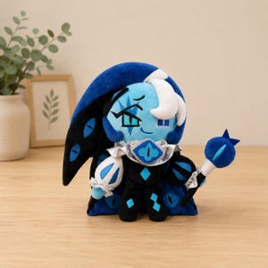 Puede incluir: Muñeco de peluche con un personaje de piel azul con una capa azul oscuro y un tocado blanco y azul. El muñeco tiene un bastón con un orbe azul y está decorado con formas de diamante y motivos de ojos. El muñeco mide aproximadamente 25 cm de alto.