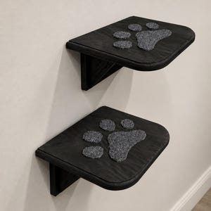 May include: Two black wall-mounted shelves with rounded front corners. Each shelf features a textured gray paw print inlay on the top surface. The shelves are constructed with a flat horizontal platform and a triangular support bracket underneath.