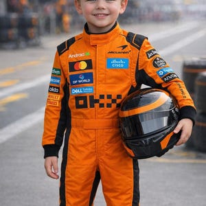 Könnte beinhalten: Ein Kind in einem orangefarbenen und schwarzen Rennanzug, das einen schwarzen und orangefarbenen Helm hält. Der Anzug hat Sponsorenlogos wie "McLaren", "Android" und "Cisco". Das Kind lächelt und posiert auf einer Rennstrecke.