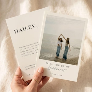 May include: A cream-colored card with the text "HAILEY," and a photograph of two women on a beach. The card reads "WILL YOU BE MY Bridesmaid?" and includes the date 08.09.2026. The card is held by a hand with gold rings.