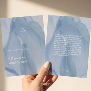 Peut inclure: Deux cartes bleues sur le thème floral avec du texte. Une carte indique "Chère Claire, je ne peux pas dire oui sans toi. Veux-tu être mon quelque chose de bleu ?" L'autre carte contient un poème. Les cartes sont tenues par une main.