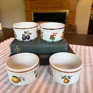 May include: Four white ceramic ramekins with vertical ribbing and a gold rim. Each dish features colorful illustrations of various fruits including plums, red currants, pears, and lemons. The ramekins are arranged in two pairs on a dark green book.