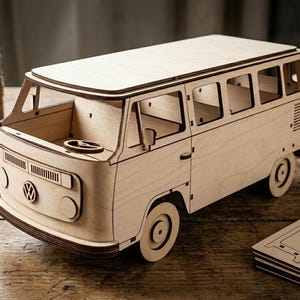 May include: A light-colored wooden model of a Volkswagen bus featuring a flat front, a steering wheel, and multiple window cutouts. The vehicle has a classic van silhouette with circular wheels and a prominent Volkswagen logo on the front panel.