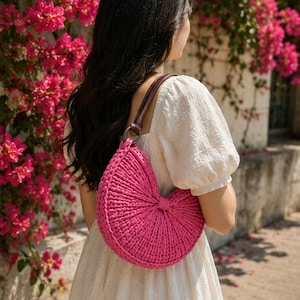 May include: A semi-circular hot pink crochet handbag featuring a textured, ribbed knit pattern and a dark brown leather shoulder strap attached with a circular metal ring. The bag has a distinct pleated design that radiates from the center point.