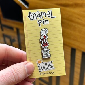 May include: A yellow card with the text "enamel pin" written in black. A red and white enamel pin of a lobster in a bucket with the text "Escape Fate" is attached to the card. The card also has the text "Fort House Studios" and "@forthouse" in black.