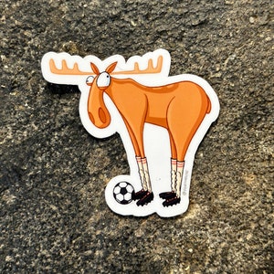 May include: A cartoon sticker of an orange moose wearing black and white striped socks and black soccer cleats. The moose is looking to the left and has a surprised expression. The moose is holding a black and white soccer ball with its right foot. The sticker has the text "@forthelove" in the bottom right corner.