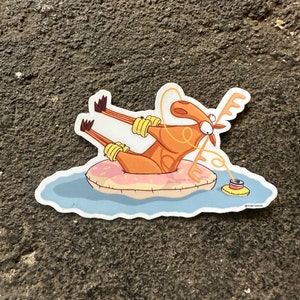 Peut inclure: Un élan de dessin animé portant des brassards jaunes flotte sur une bouée gonflable rose et blanche dans une eau bleue. L'élan tient une ficelle attachée à une canette rouge et jaune.