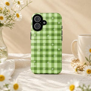 Könnte beinhalten: Eine grüne Handyhülle mit Vichy-Muster und kleinen weißen Blumenakzenten. Die Hülle liegt auf einer weißen Oberfläche mit einer Vase mit Gänseblümchen und einer Tasse mit Gänseblümchenmuster.