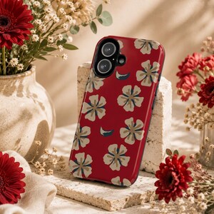 Könnte beinhalten: Eine rote Handyhülle mit einem Blumenmuster aus cremefarbenen und blauen Blüten. Die Hülle wird auf einem Steinblock präsentiert, mit roten Gerbera-Gänseblümchen und anderen Blumen im Hintergrund.