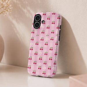Könnte beinhalten: Eine Smartphone-Hülle mit einem hellrosa-weißen Vichy-Karo-Muster. Das Design ist mit einem sich wiederholenden Aufdruck aus kleinen roten Kirschen mit grünen Stielen und winzigen weißen Blüten verziert, die über die Oberfläche verteilt sind.