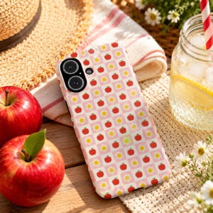 Könnte beinhalten: Eine Smartphone-Hülle mit einem sich wiederholenden Schachbrettmuster aus hellrosa und weißen Quadraten. Das Design zeigt kleine rote Apfelillustrationen und gelbe Blumensymbole, die in abwechselnden Quadraten zentriert sind.